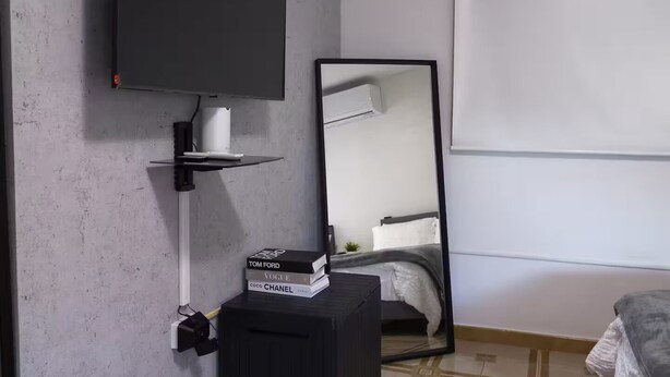 Ein Schlafzimmer mit flachbildfernseher an der Wand, einem Nachttisch mit Spiegel, einem Bett mit grauer Decke und einem gemusterten Boden.