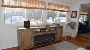 A living room with a wooden cabinet, a fireplace, and a view of a snowy landscape through large windows with bamboo blinds. - Waterfront 1860s Farmhouse, Private Hot Tub, Fire Pit, Pond, Canoe, Walk to Town (Chestertown)
