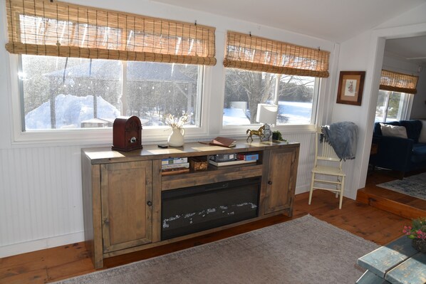 A living room with a wooden cabinet, a fireplace, and a view of a snowy landscape through large windows with bamboo blinds.