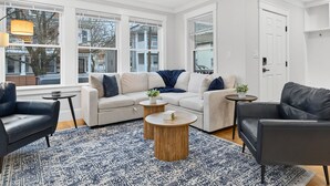 A modern living room with a beige sofa, a black leather armchair, and a round wooden coffee table. Large windows provide natural light and a view of the outside. - Somerville 4BR: 2 Parking/Yard/Game Rm—Near Boston (Somerville)