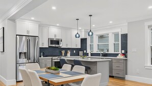 A modern kitchen with white cabinets, stainless steel appliances, a central island with seating, and pendant lighting. - Somerville 4BR: 2 Parking/Yard/Game Rm—Near Boston (Somerville)