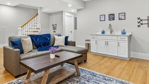 A modern living room with a sofa, coffee table, and a kitchen area with cabinets and a sink. - Somerville 4BR: 2 Parking/Yard/Game Rm—Near Boston (Somerville)
