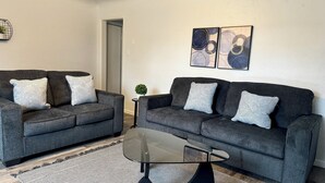 A living room with two grey sofas, each with patterned cushions, a glass coffee table, a small side table with a plant, and abstract wall art.