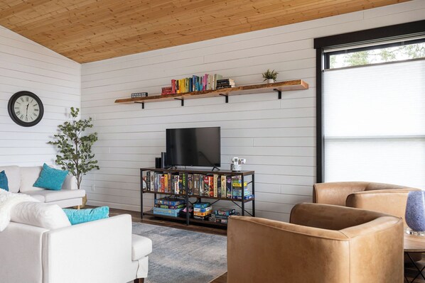 A modern living room with a white couch, a coffee table, a bookshelf, a TV, and a window with blinds.