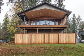 A two-story wooden cabin with a balcony, surrounded by trees and a wooden fence. - The Treasure Trove - A Dog-Friendly Waterview Gem (Grapeview)