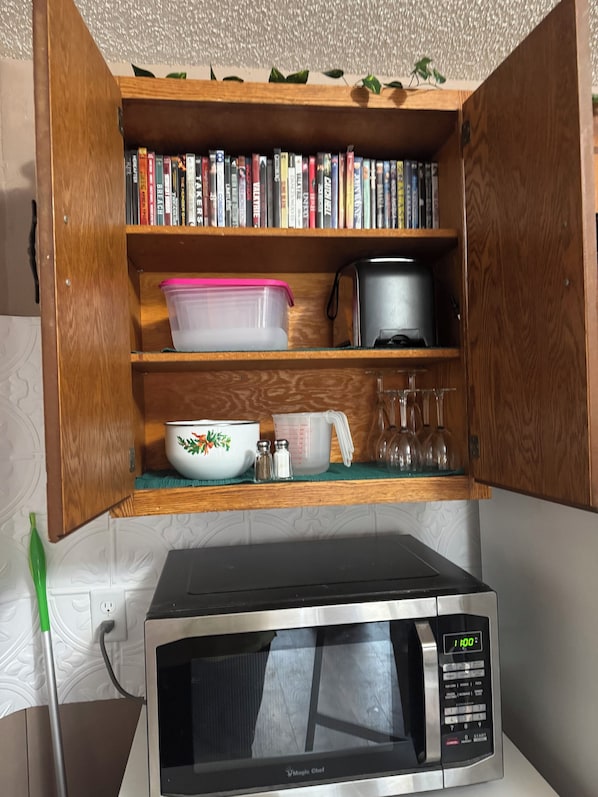 Una cocina con microondas, un mueble de madera con libros, un recipiente de plástico, un cuenco decorativo, saleros y pimenteros, una taza medidora y una planta verde en el estante.