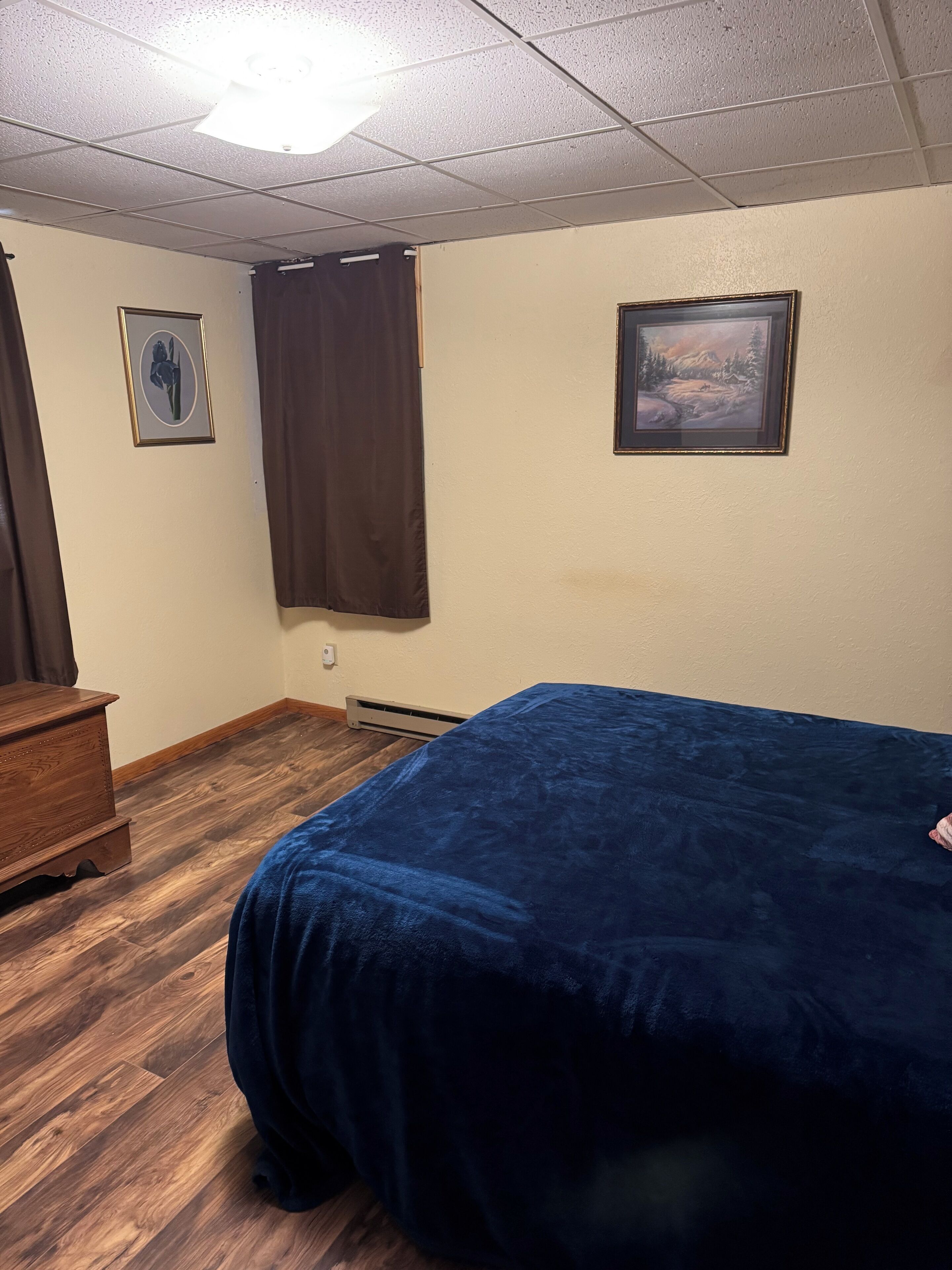 A room with a bed, a dresser, a window with curtains, and two framed pictures on the wall.