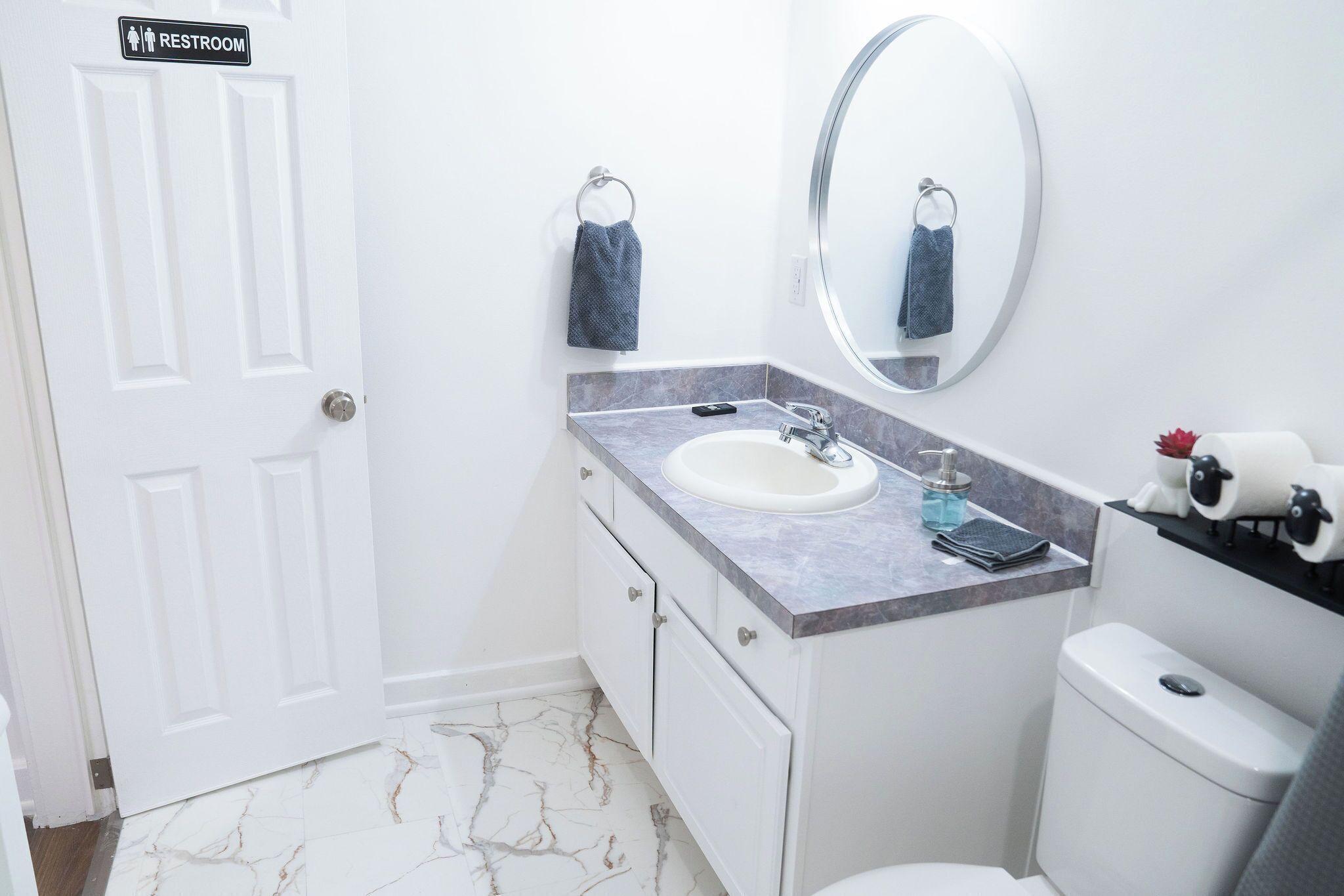 A bathroom with a white door marked "RESTROOM," a marble-tiled floor, a white sink with a soap dispenser, and a toilet.