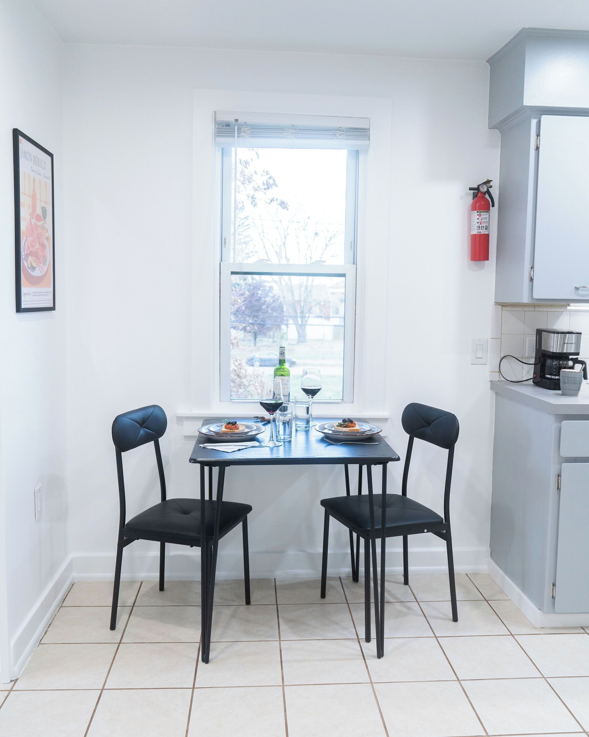 A small, bright kitchen with a dining table set for two, a window with a view of trees, and a fire extinguisher on the wall.