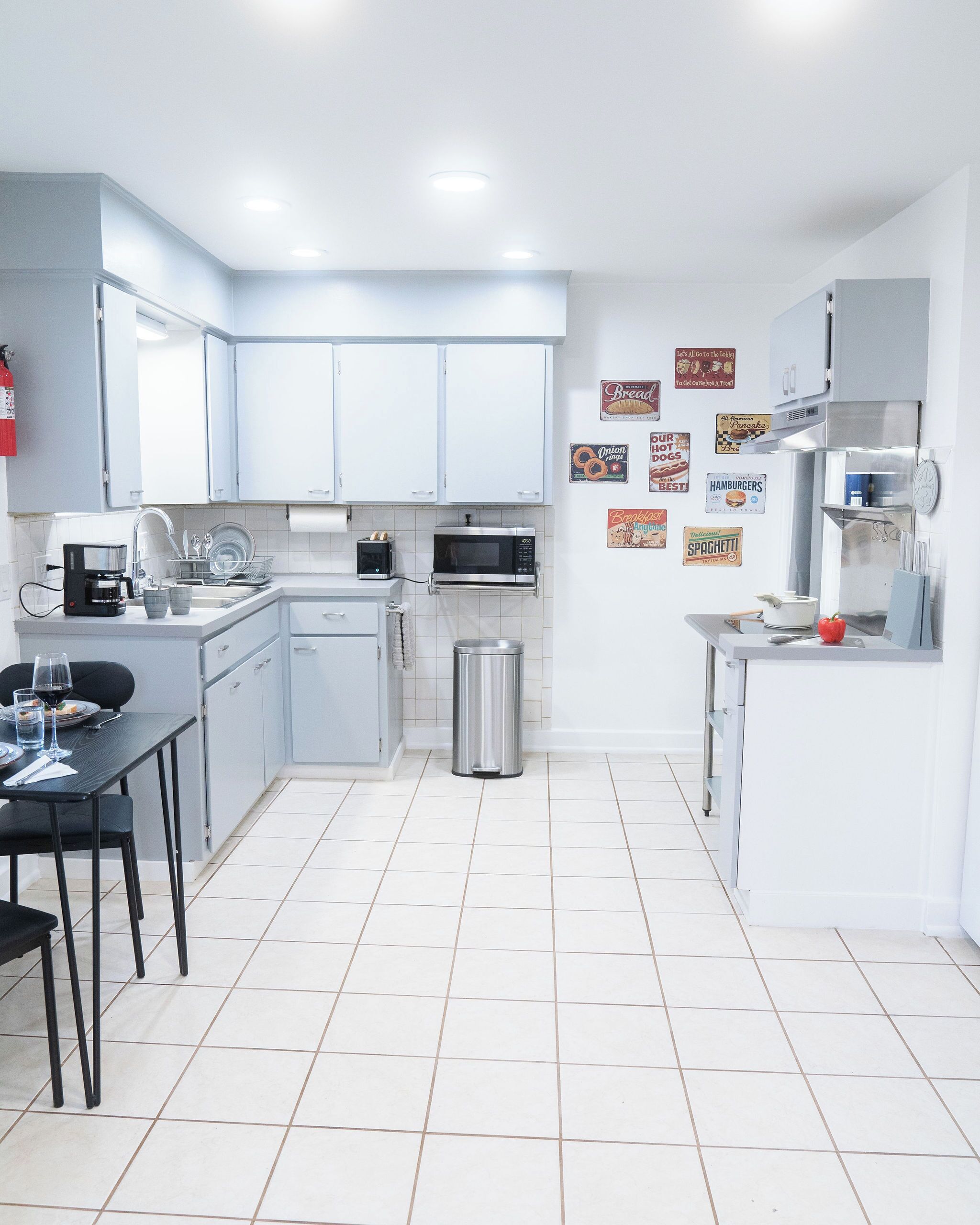 A modern kitchen with white cabinets, a sink, a microwave, and a trash can. There are wall decorations and a table with chairs.