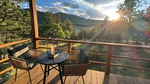 Une terrasse en bois avec une table ronde et deux chaises, offrant une vue sur un paysage montagneux au coucher du soleil.
