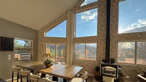 Un vaste espace dédié à la salle à manger, avec une grande table en bois et six chaises, une cheminée en pierre et de grandes fenêtres offrant une vue sur l’extérieur.