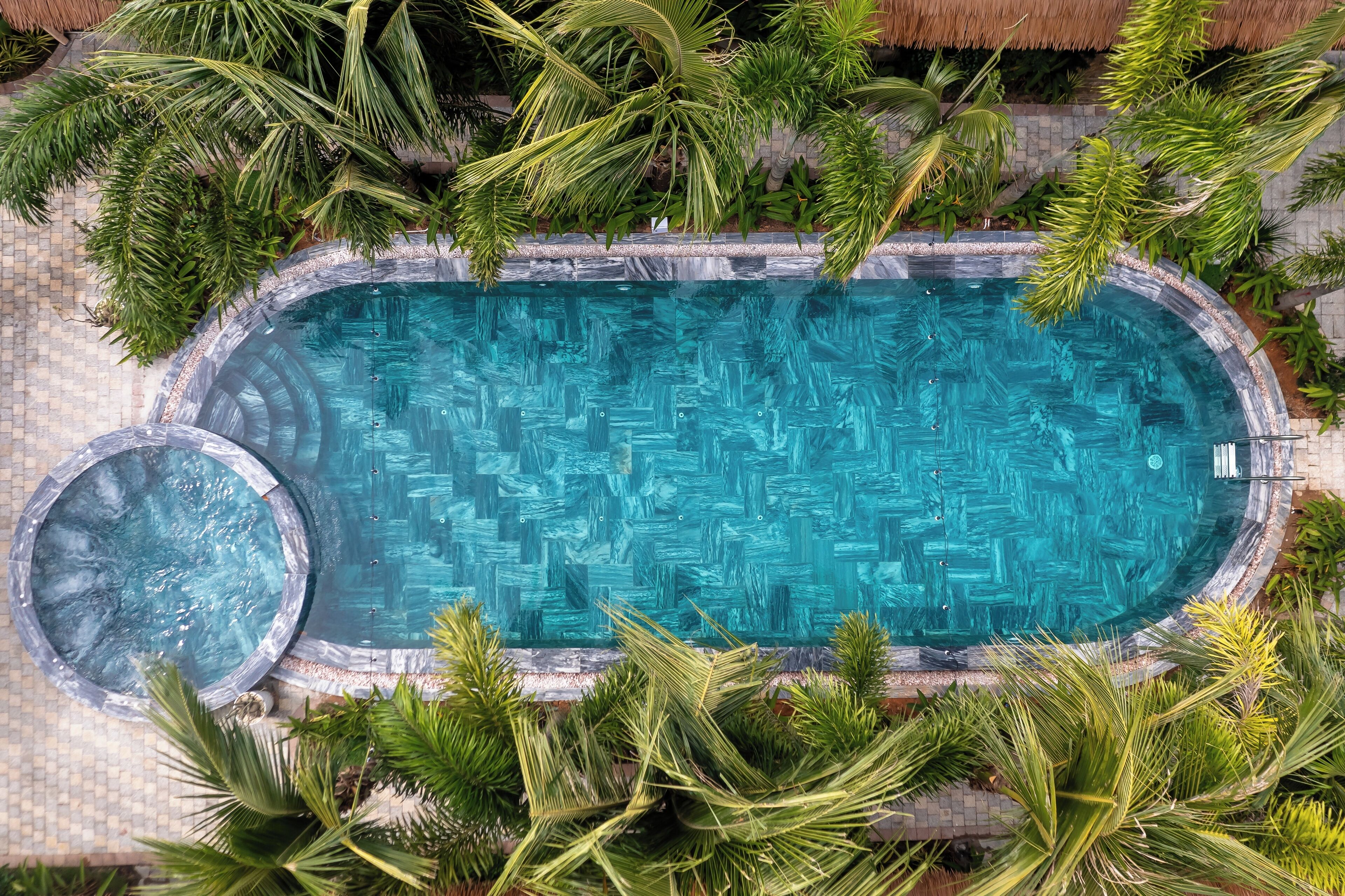 A rectangular swimming pool with a hot tub, surrounded by palm trees and a thatched roof structure.