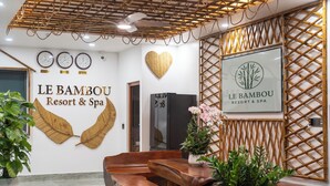 Ein Empfangsbereich mit Bambus-Dekor, darunter ein Schild für Le Bambou Resort & Spa, Holzbänke und eine wandmontierte Uhr.