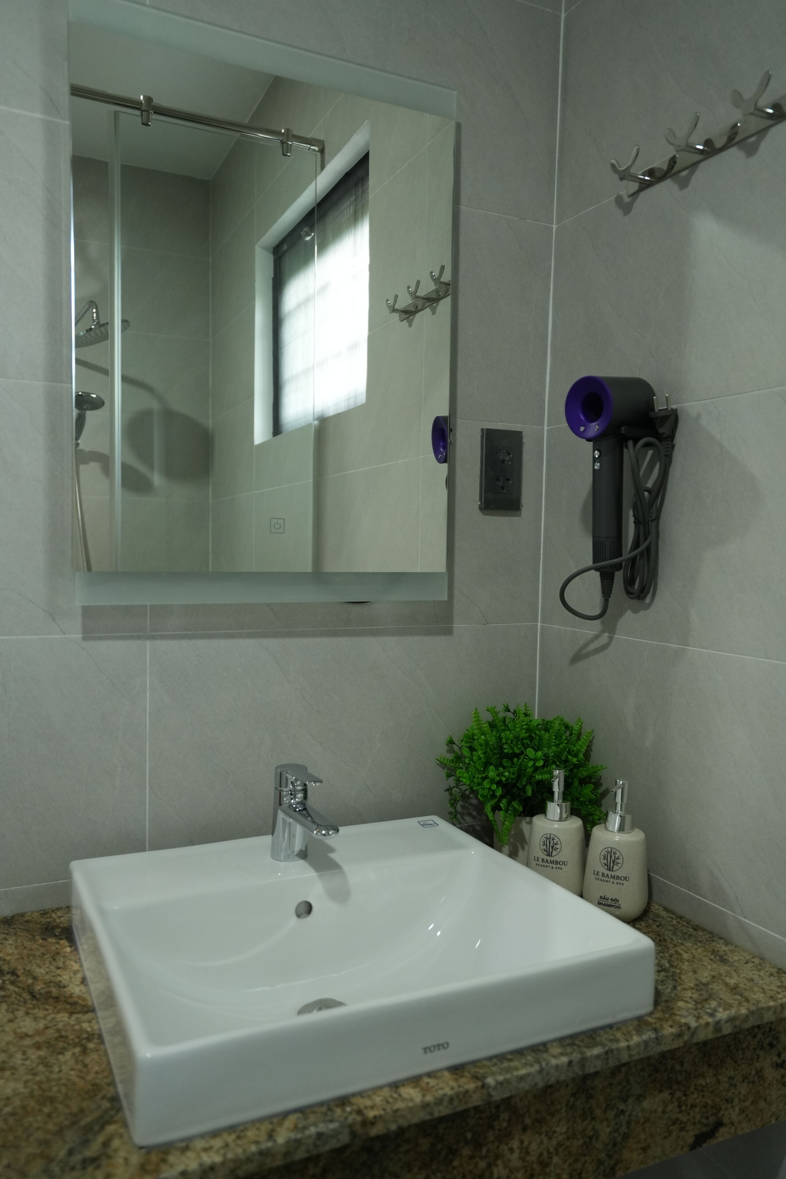 Une salle de bain avec un lavabo blanc, un miroir, un sèche-cheveux et une petite plante.