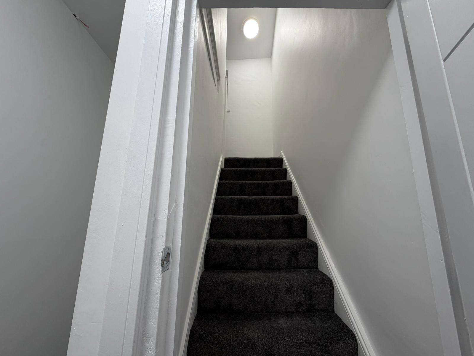 Eine enge, mit Teppich belegte Treppe, an deren oberen Ende sich eine kleine, runde Leuchte an weißen Wänden befindet.