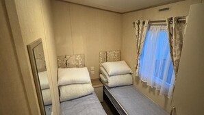 Una stanza piccola e accogliente con due letti singoli, una lampada a soffitto, una finestra con tende trasparenti e uno specchio appeso al muro.