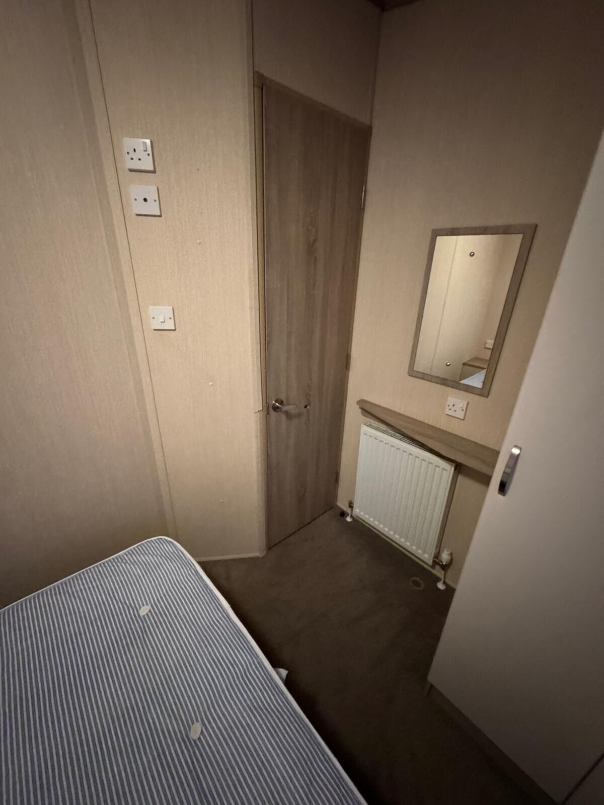 A small, narrow room with a single bed, a door, a radiator, and a mirror on the wall.