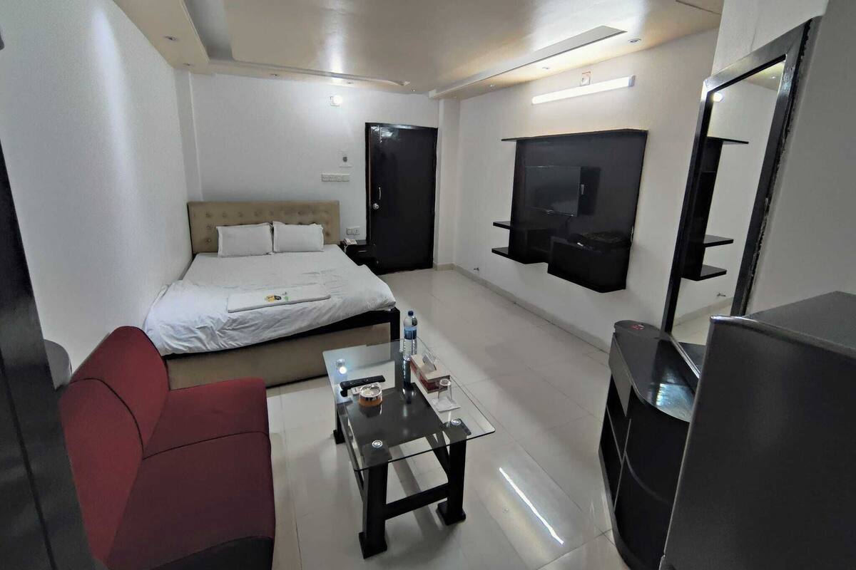 A hotel room with a bed, a red chair, a glass coffee table, a TV mounted on the wall, and a mirror.