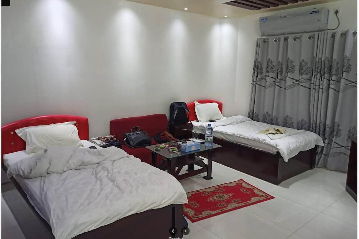 A hotel room with two single beds, a small table with a water bottle, a red bag, and a red chair.