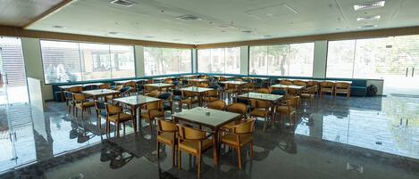 A spacious dining area with multiple tables and chairs, large windows, and a high ceiling with recessed lighting.