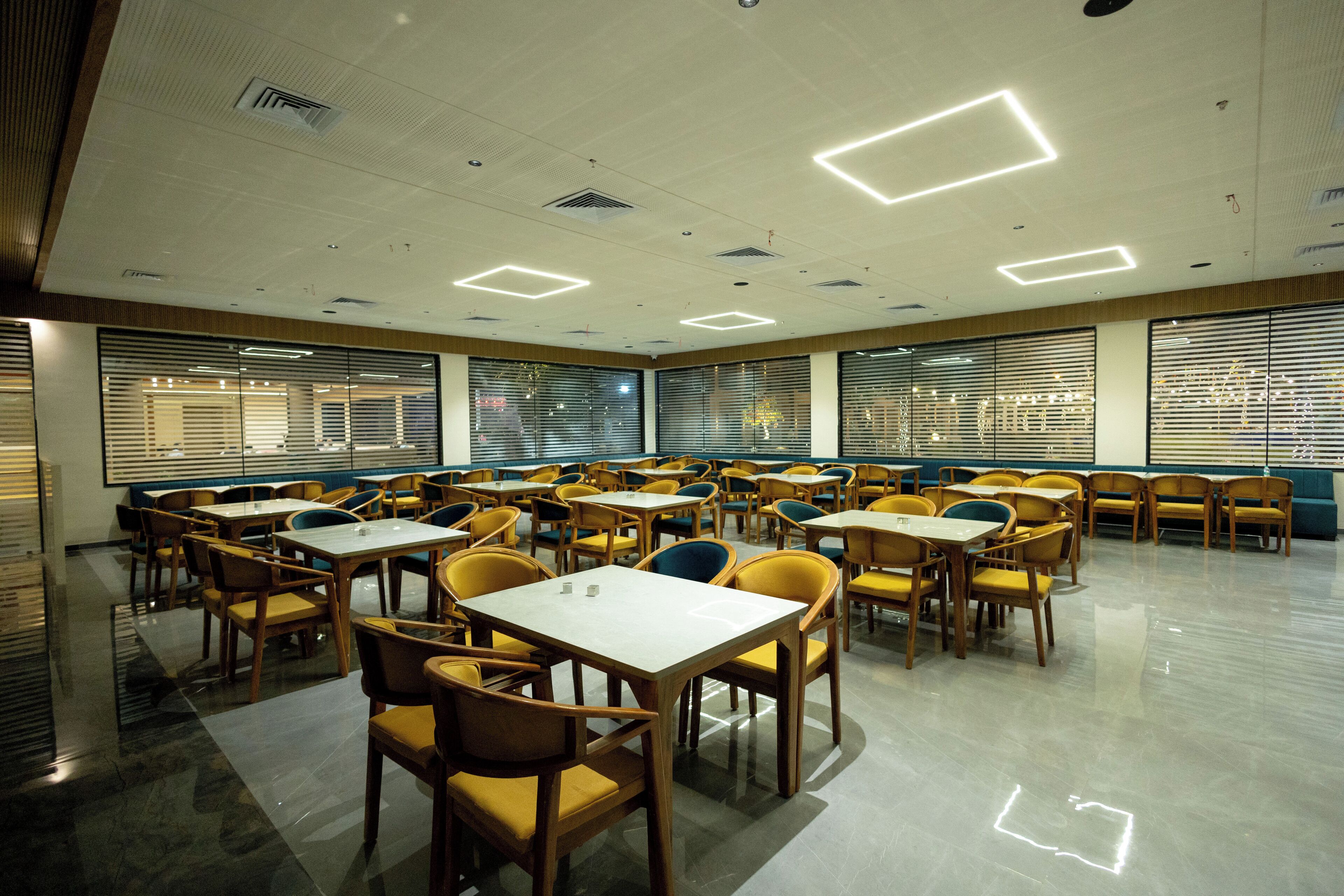 A spacious dining area with multiple tables and chairs, large windows with blinds, and a clean, modern ceiling with recessed lighting.