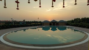 A large, circular swimming pool with clear water reflecting the sky. Surrounding the pool are low-rise buildings with flat roofs, likely part of a resort or hotel. - Madhuvan Resort (Talala)