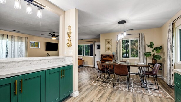 A spacious kitchen with green cabinets, a dining area with a wooden table and chairs, and a large window with curtains. - Buckthorn Beauty: Private Hot Tub & Lounge Space (Pine Valley)