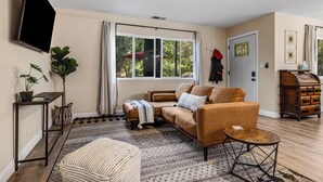 A living room with a sofa, a coffee table, a TV, and a large window with curtains. - Buckthorn Beauty: Private Hot Tub & Lounge Space (Pine Valley)