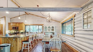 Une cabane en rondins dotée d’une cuisine avec des meubles en bois, d’un coin repas avec une table et des chaises, et de grandes fenêtres laissant entrer la lumière naturelle.