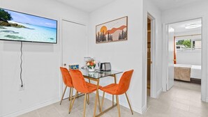 A modern dining area with a glass table and orange chairs, a flat-screen TV on the wall, and a painting above the table. - Beautiful 1-bedroom bungalow with AC in pleasant Tustin (Tustin)