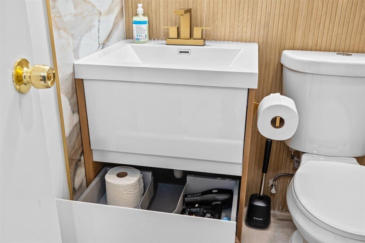 A bathroom with a white sink, toilet, and a hidden storage compartment with a toilet paper roll and cleaning supplies.