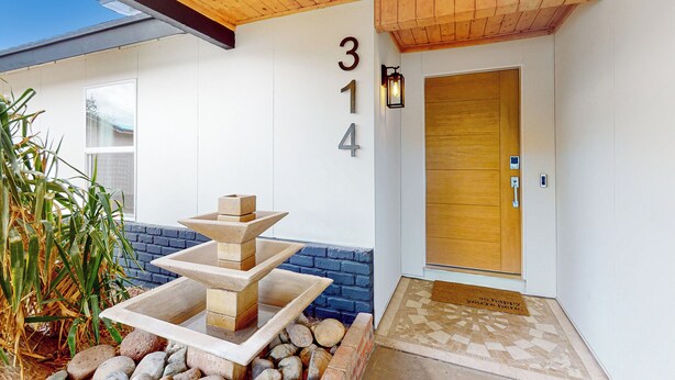 Ein moderner Gebäudeeingang mit Holztür, einem Schild mit der Nummer 314, einem kleinen Wasserspiel im Freien und einer Fußmatte.