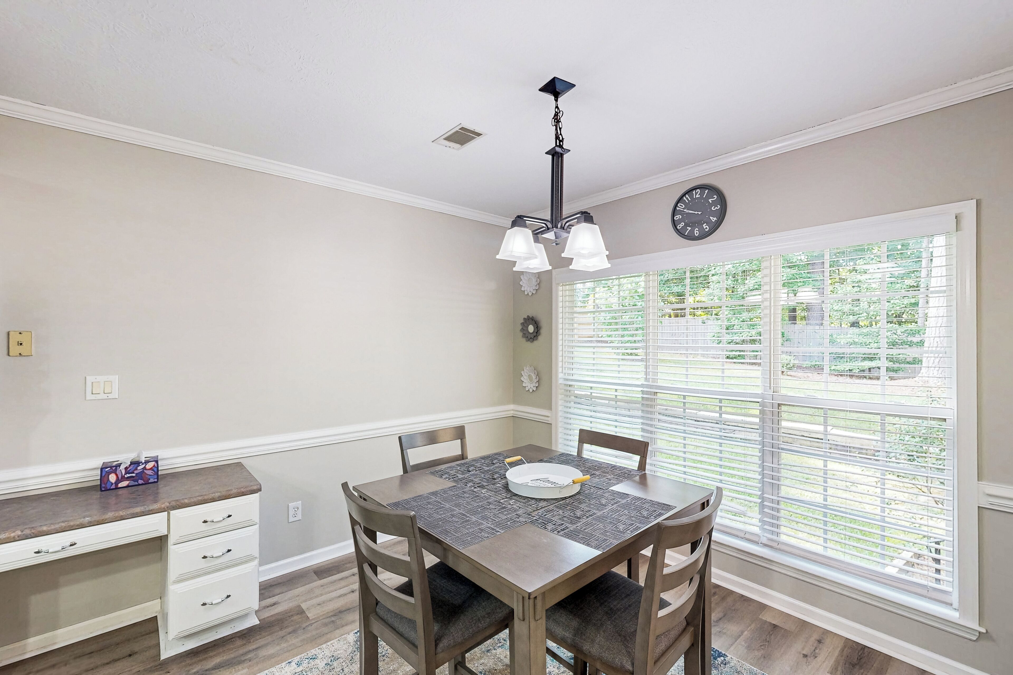 A dining area with a table, chairs, a chandelier, a clock, and a window with blinds.