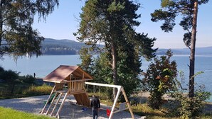 Una casita de juegos de madera con un columpio y un tobogán, rodeada de árboles y vista a un lago.