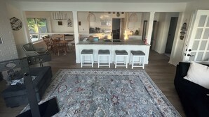 Un vaste salon agrémenté d’un grand tapis, un coin repas avec des chaises, une cuisine dotée d’un îlot central et de tabourets de bar, et une porte avec une pancarte.