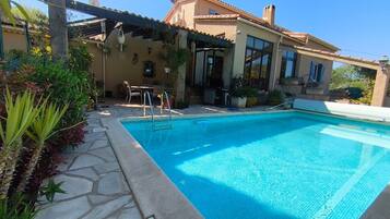 Une piscine avec une terrasse en pierre, une pergola ornée de plantes suspendues et un bâtiment doté d’un toit en tuiles.