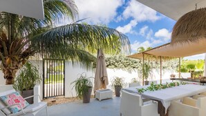 Un patio al aire libre con una mesa y sillas blancas, rodeado de plantas en macetas y una palmera.