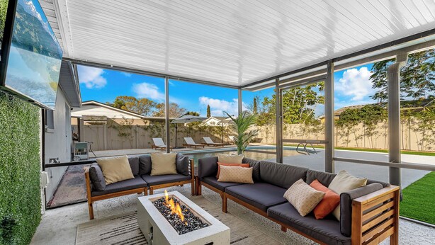 Ein moderner Außenbereich mit Feuerstelle, zwei Sofas und einem großen Panoramafenster mit Blick auf einen Pool und Grünflächen.