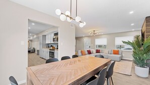 Eine moderne Küche mit weißen Schränken, ein Essbereich mit einem Holztisch im Fischgrätmuster und eine Wohnbereich mit einem grauen Sofa und orangefarbenen Kissen.