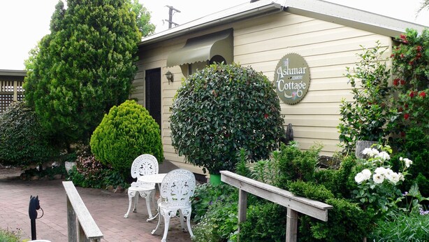 A cottage with a sign reading "Ashmar Cottage," surrounded by lush greenery and a paved patio area with white metal chairs. - Ashmar offers  you a  true country lifestyle.. (Gloucester)