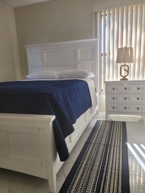 Un dormitorio con cama blanca, cubrecama azul oscuro, mesita de noche blanca con cajones y una lámpara.