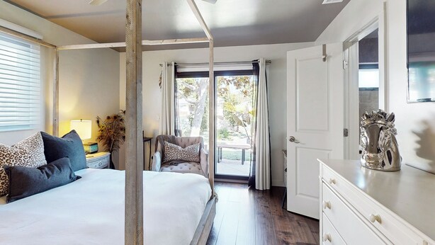 Un dormitorio con una cama grande, una mesita de noche con lámpara, una silla y vistas al exterior a través de un ventanal.