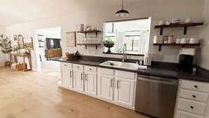 A modern kitchen with white cabinets, a stainless steel dishwasher, and a black countertop. There are shelves above the sink holding cups and a potted plant. - Hot Tub & Game Room! Pinetop 'Wild Horse Retreat' (Pinetop)