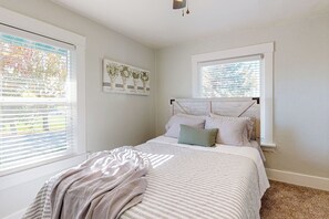 Un dormitorio con una cama, dos ventanas con persianas, un cabecero y una obra de arte colgada en la pared.