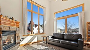 Een gezellige woonkamer met een open haard, een leren bank en grote ramen met uitzicht op een huis en bergen.