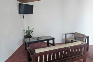 Una habitación con un banco de madera, una pequeña mesa, una silla, una planta en maceta y un televisor montado.