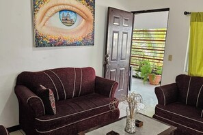 A living room with a dark red sofa, a painting of an eye on the wall, and a door leading to a balcony with plants. - Tierra Viva Managua - Villa of rest and nature (Managua)