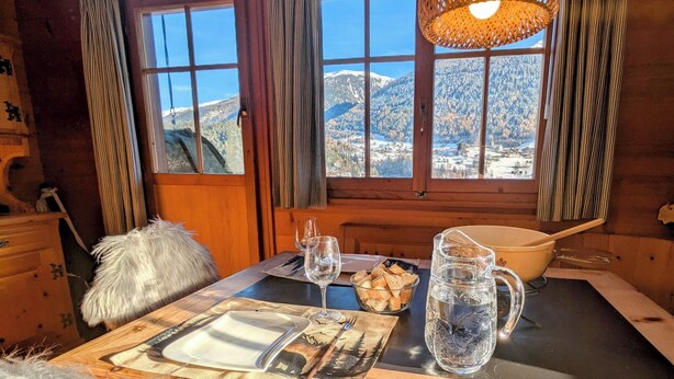 Un acogedor cuarto de cabaña de madera con una mesa dispuesta para el desayuno, vista a montañas nevadas y un ventanal con cortinas.