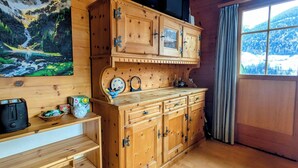 Interior de una cabaña de madera con un gran armario de madera, un cuadro de un paisaje montañoso y una ventana con vistas a colinas nevadas.